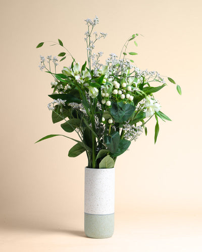 Die Vase Luftkuss mit ihrem modernen, weiß und hellgrün gesprenkelten Design hält einen Strauß aus grünen Blättern, weißen Beeren und zarten kleinen weißen Blüten vor einem einfarbig beigen Hintergrund.