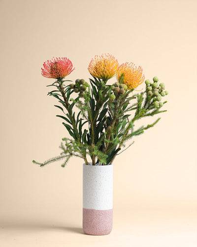 Die Couture-Keramikvase mit rosa-weißem Farbverlauf enthält einen leuchtenden feurigen Farben-Blumenstrauß mit drei Nadelkissen-Proteen und grünem Blattwerk, alles vor einem schlichten beigen Hintergrund.