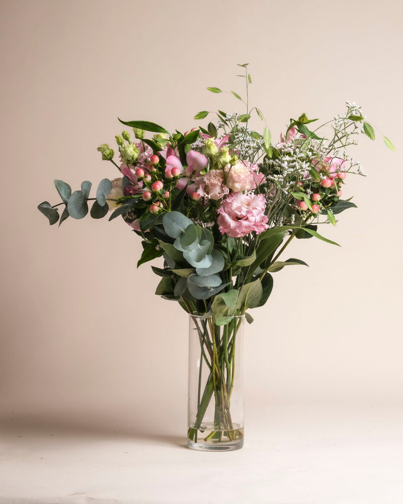 In einer klaren Glasvase steht der Ballerina-Strauß mit rosa Blumen, Eukalyptusblättern und Grünzeug vor einem schlichten beigen Hintergrund.