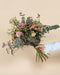 Eine Hand in einem weißen Rüschenärmel hält den "Morgenlicht"-Strauß aus gemischten Wildblumen und Grünzeug mit rosa, pfirsichfarbenen und cremefarbenen Herbstblüten vor einem schlichten beigen Hintergrund.