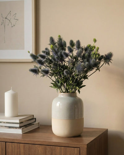 Eine Edeldistel mit blauen Stachelblüten in einer Keramikvase steht auf einem Holzschrank neben einer weißen Kerze und gestapelten Büchern, dahinter an der beigen Wand eine gerahmte minimalistische Strichzeichnung.