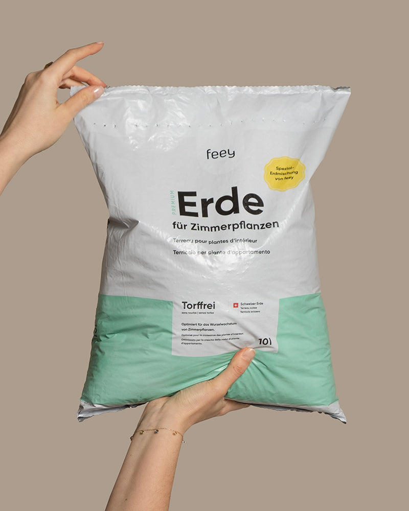 Zwei Hände halten einen mintgrün-weißen „Feey“-Beutel mit der Aufschrift „Erde für Zimmerpflanzen“ und „Torfrei“, genehmigt von Pflanzendoktor Janko.