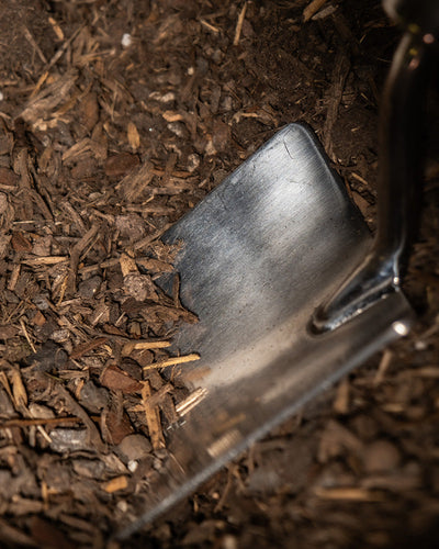 Nahaufnahme einer Metallschaufel, die in Erde gräbt, die mit kleinen Holzspänen und organischem Abfall vermischt ist. Die Kante der Schaufel ist leicht abgenutzt, was auf häufigen Gebrauch hindeutet. Die Erde für Balkon- & Gartenpflanzen um die Schaufel herum erscheint trocken und locker, perfekt für den Pflanzenanbau in Ihrem Garten oder auf Ihrem Balkon.