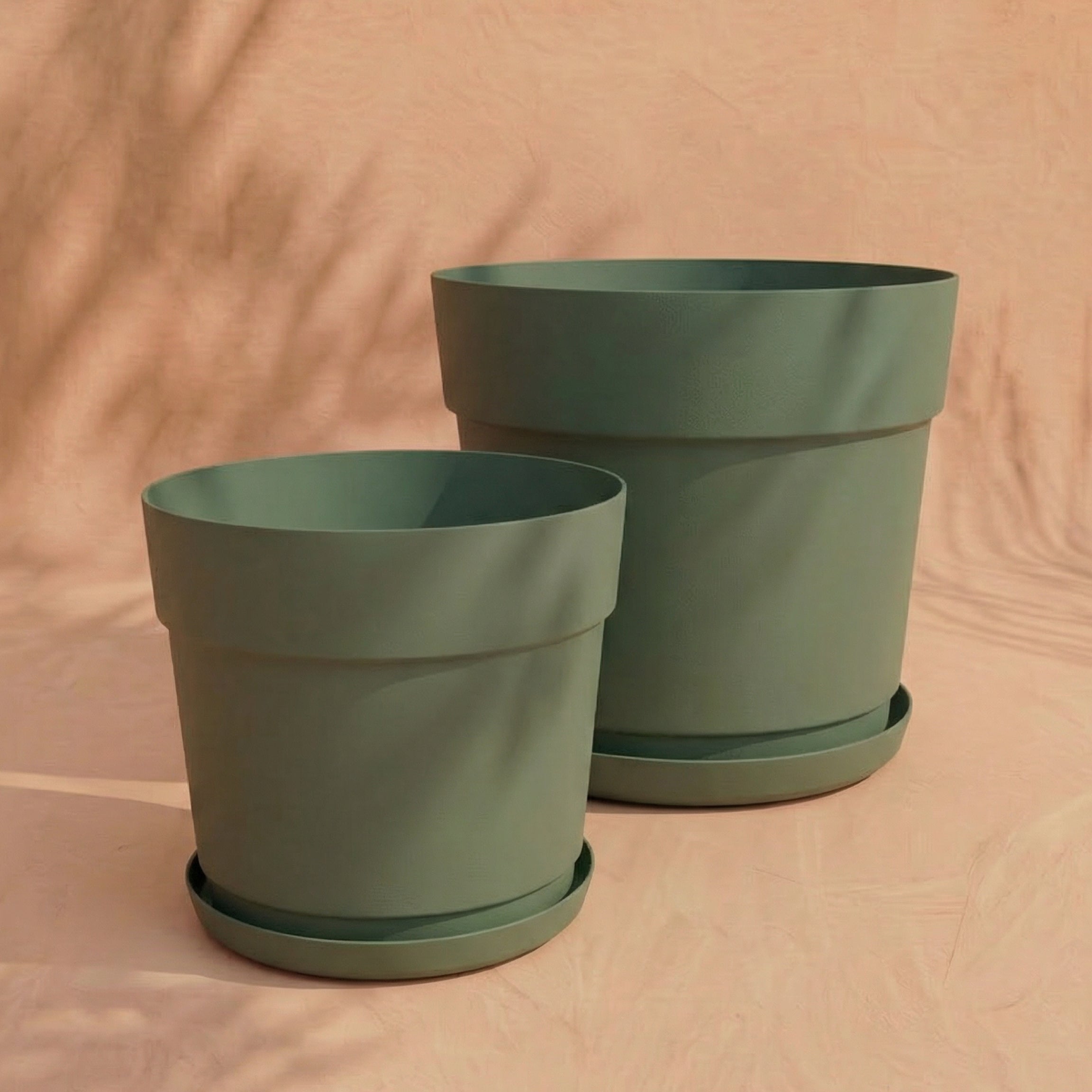 Zwei leere grüne Pflanzkübel-Set 'Bloom' (38, 29) aus recyceltem Kunststoff in zwei Größen stehen nebeneinander auf einer hell pfirsichfarbenen, strukturierten Oberfläche.