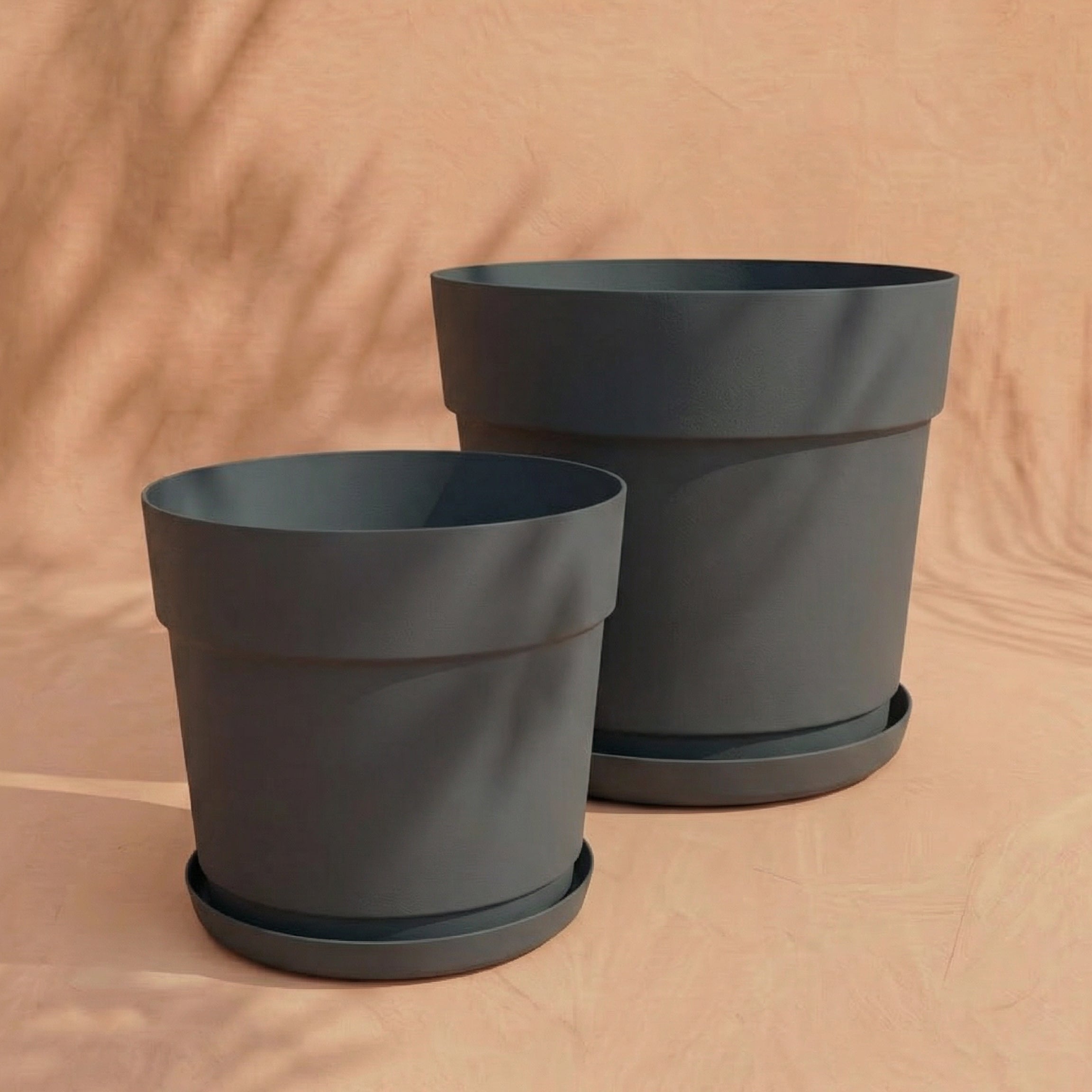 Zwei leere Pflanzkübel-Set 'Bloom' (38, 29) in mattem Grau mit passenden Untertassen aus recyceltem Kunststoff ruhen auf einer hellbeigen Fläche vor einem weichen, neutralen Hintergrund mit sanften Schatten.