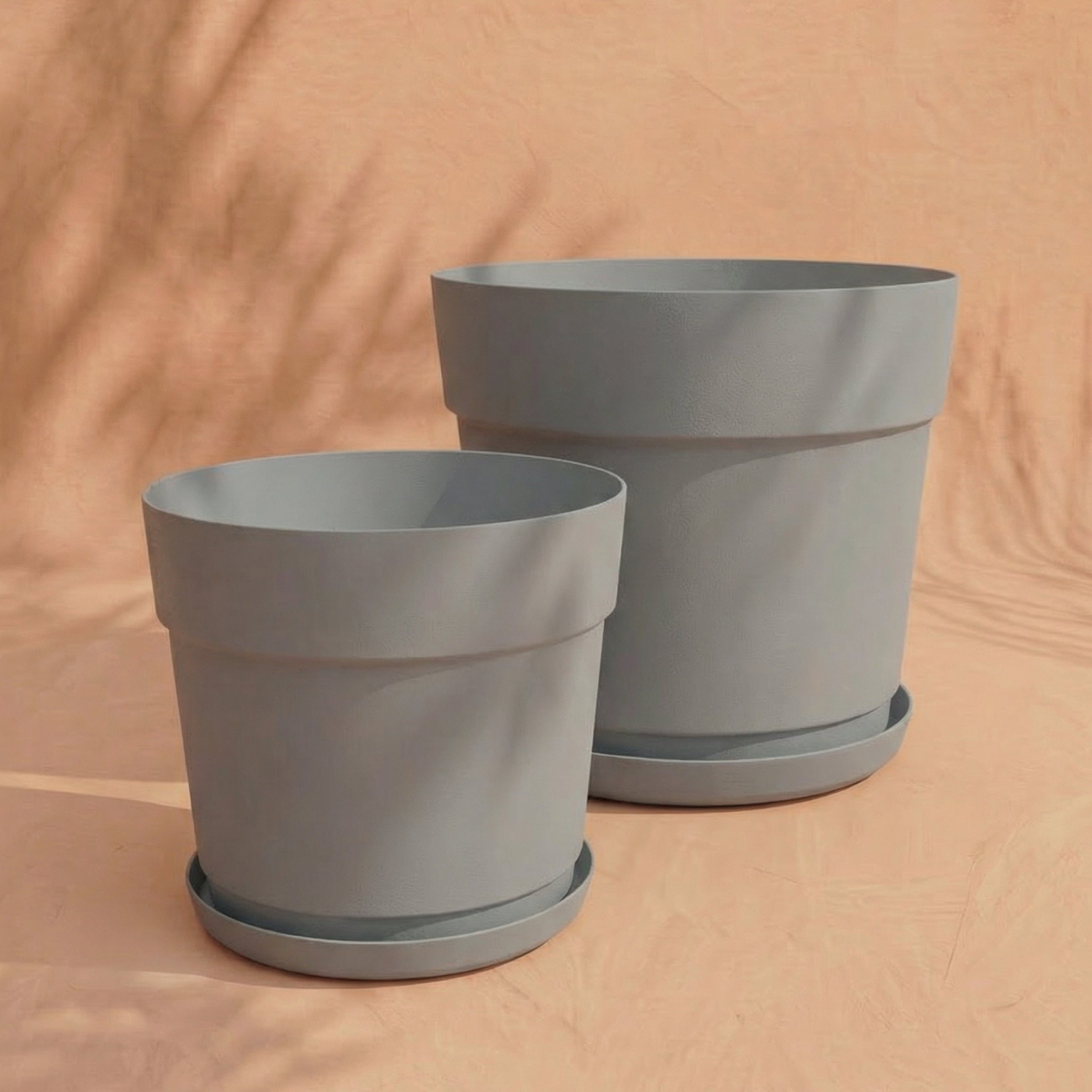 Zwei leere, hellgraue Pflanzkübel aus recyceltem Kunststoff des Sets 'Bloom' (38, 29) mit passenden Untersetzern stehen nebeneinander auf einer pfirsichfarbenen Fläche, umgeben von sanften Schattenmustern im Hintergrund.