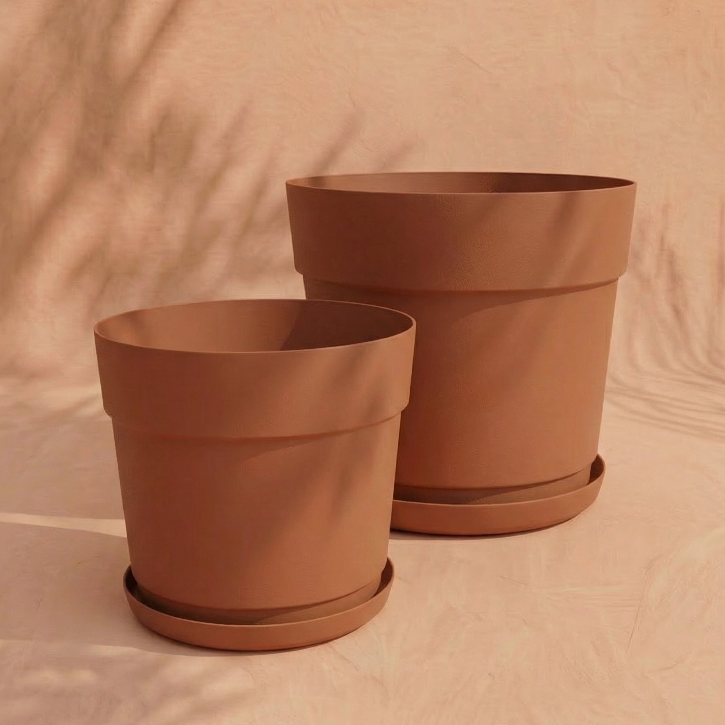 Zwei leere, UV-beständige Pflanzkübel aus dem Set 'Bloom' (38, 29) mit passenden Untersetzern stehen nebeneinander auf einer hellbeigen Fläche vor neutralem Hintergrund.