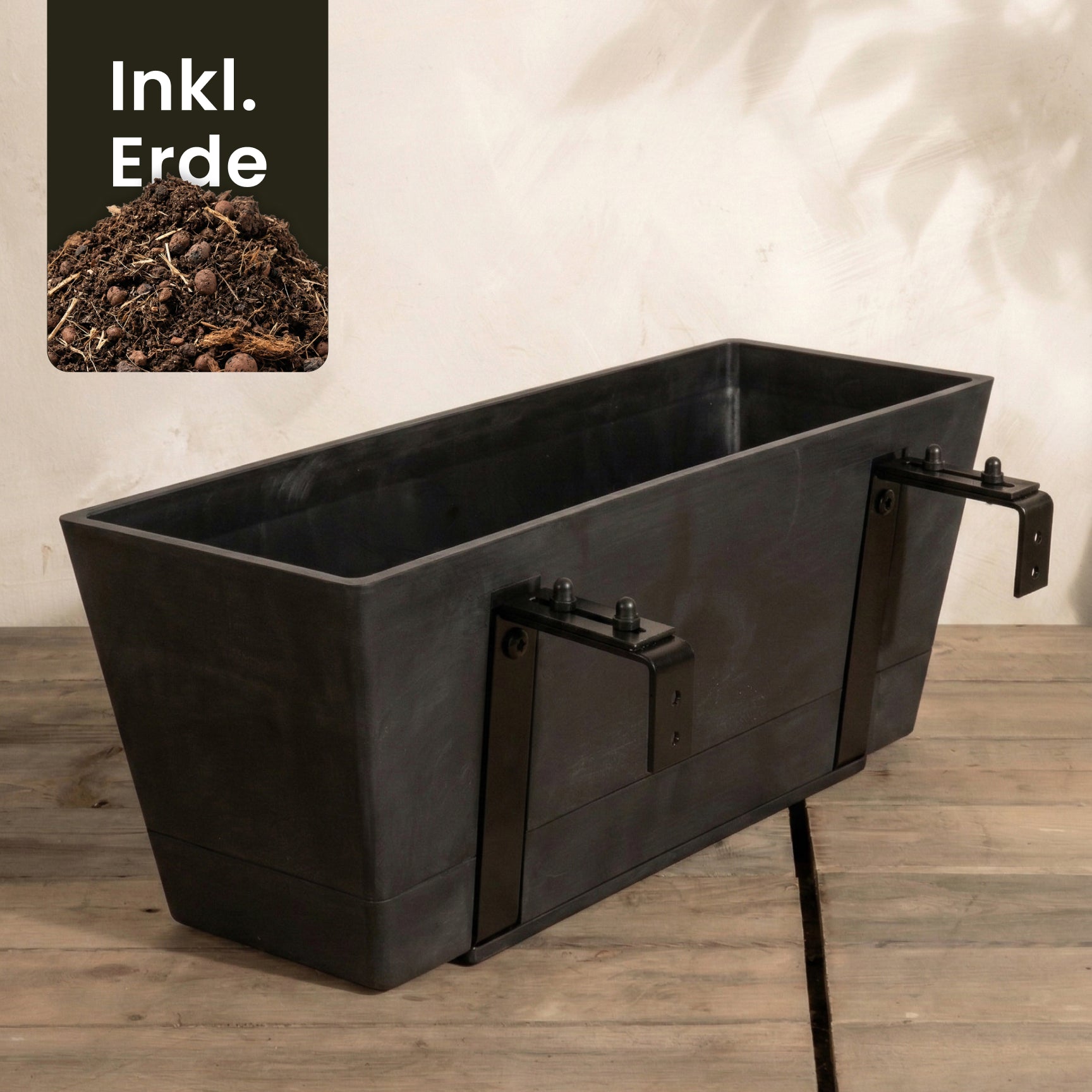 Der rechteckige schwarze Pflanzkasten für Geländer (Brügge | 48 cm) mit Hängesystem und verstellbaren Halterungen steht auf Holz. In einer Ecke sieht man Erde und den Hinweis 