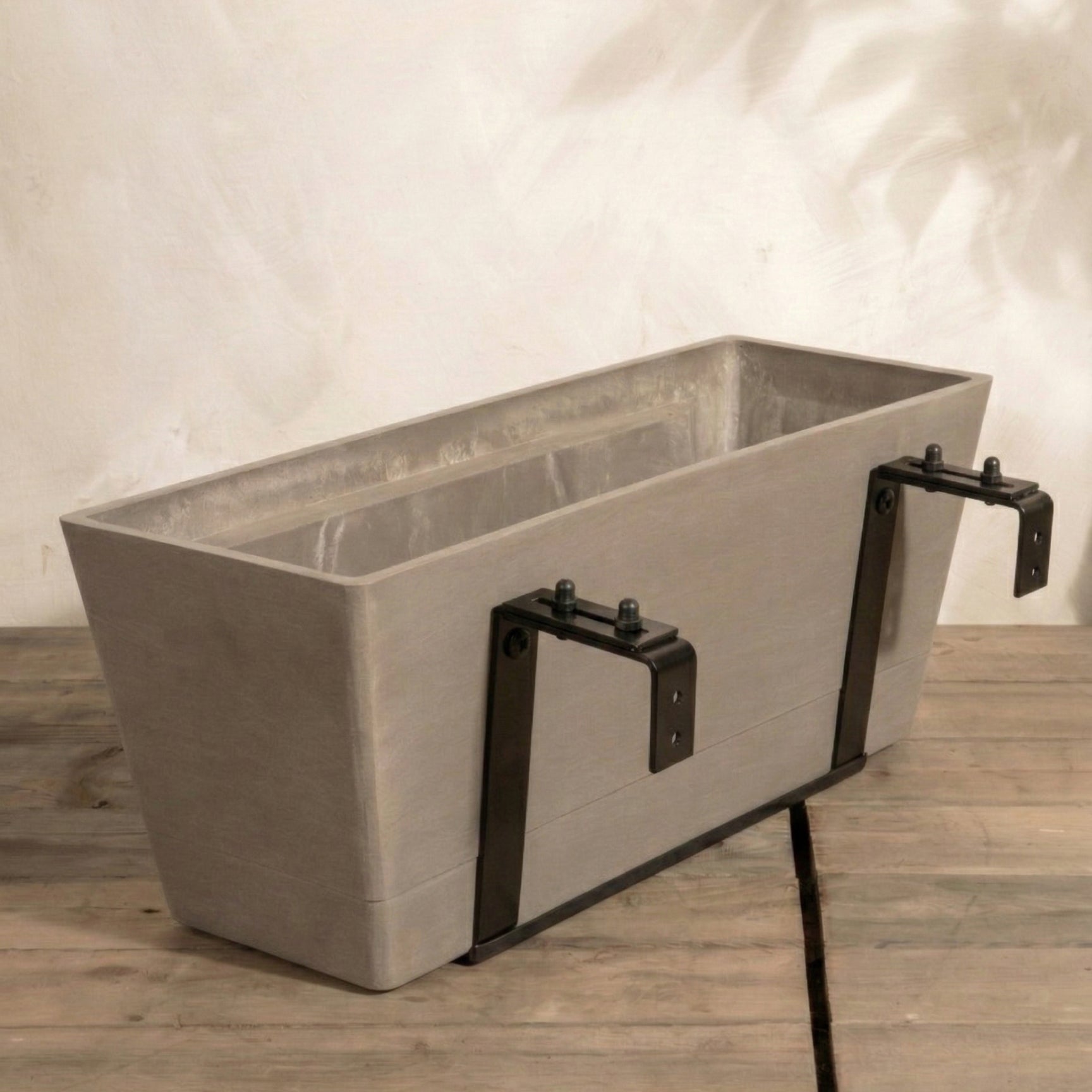 Ein grauer Pflanzkasten für Geländer (Brügge | 48 cm) mit zwei schwarzen Metallbügeln wird auf Holz vor eine helle Wand gesetzt - ideal als Beet zum Aufhängen für Ihr Hängesystem Balkon.