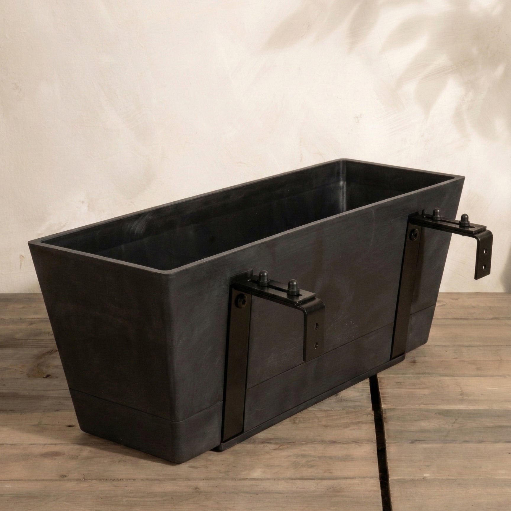 Der Pflanzkasten für Geländer (Brügge | 48 cm), ein rechteckiger schwarzer Pflanzkübel mit Metallbügeln für die Geländerbefestigung, steht auf einer Holzfläche vor einer hellen Wand.