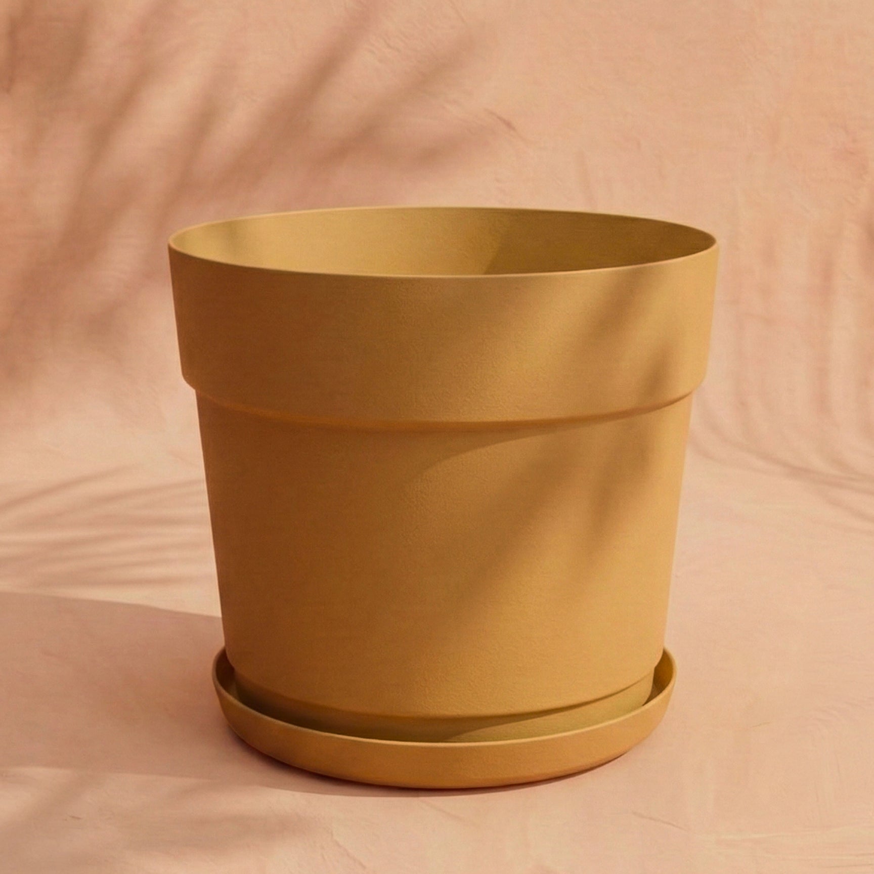 Ein großer, leerer Kunststoff-Pflanzkübel (Bloom | 29 cm Ø) in Terrakotta-Farbe mit passender Untertasse ruht auf einer hellbeigen Fläche und wirft weiche Schatten vor einem pfirsichfarbenen Hintergrund.