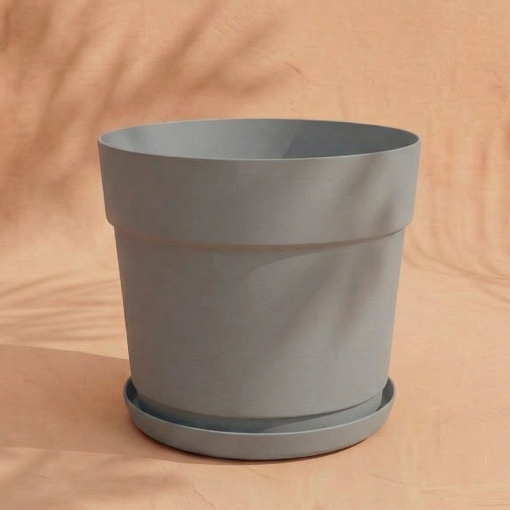 Ein hellgrauer Kunststoff Pflanzkübel (Bloom | 29 cm Ø) aus recyceltem Kunststoff mit passender Untertasse steht auf einer pfirsichfarbenen Fläche und wirft weiche Schatten in den Hintergrund.