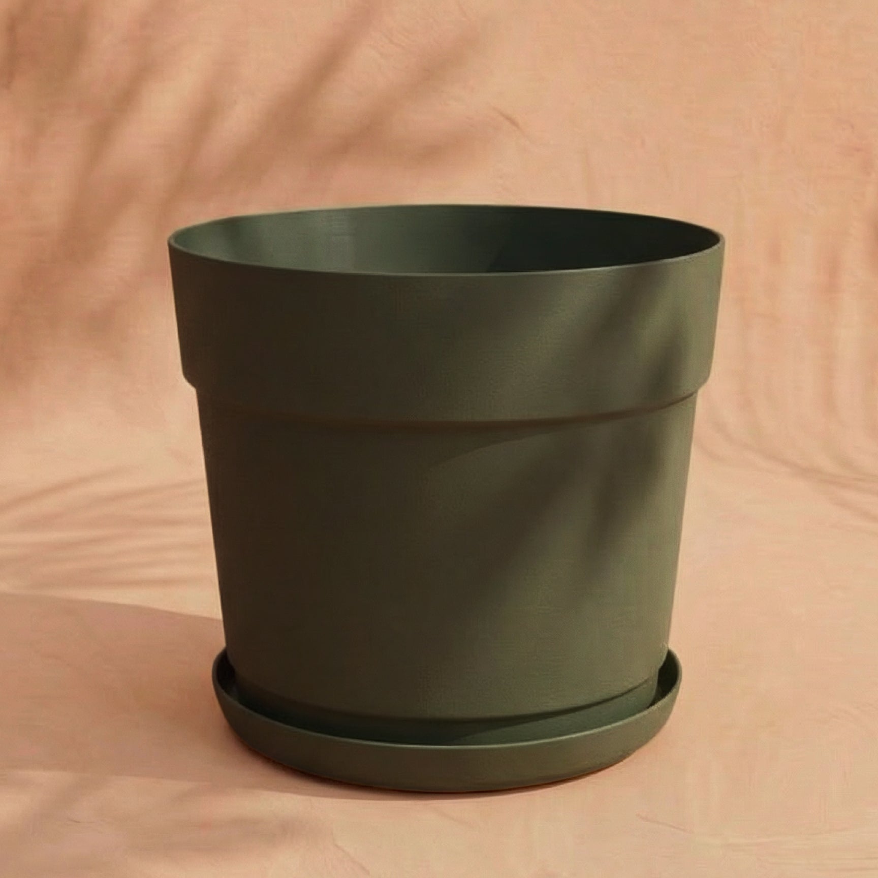 Ein schlichter, leerer Kunststoff Pflanzkübel (Bloom | 29 cm Ø) in Dunkelgrün mit passendem Untersetzer steht auf einer hellbeigen Fläche mit dezentem Schatten im Hintergrund.