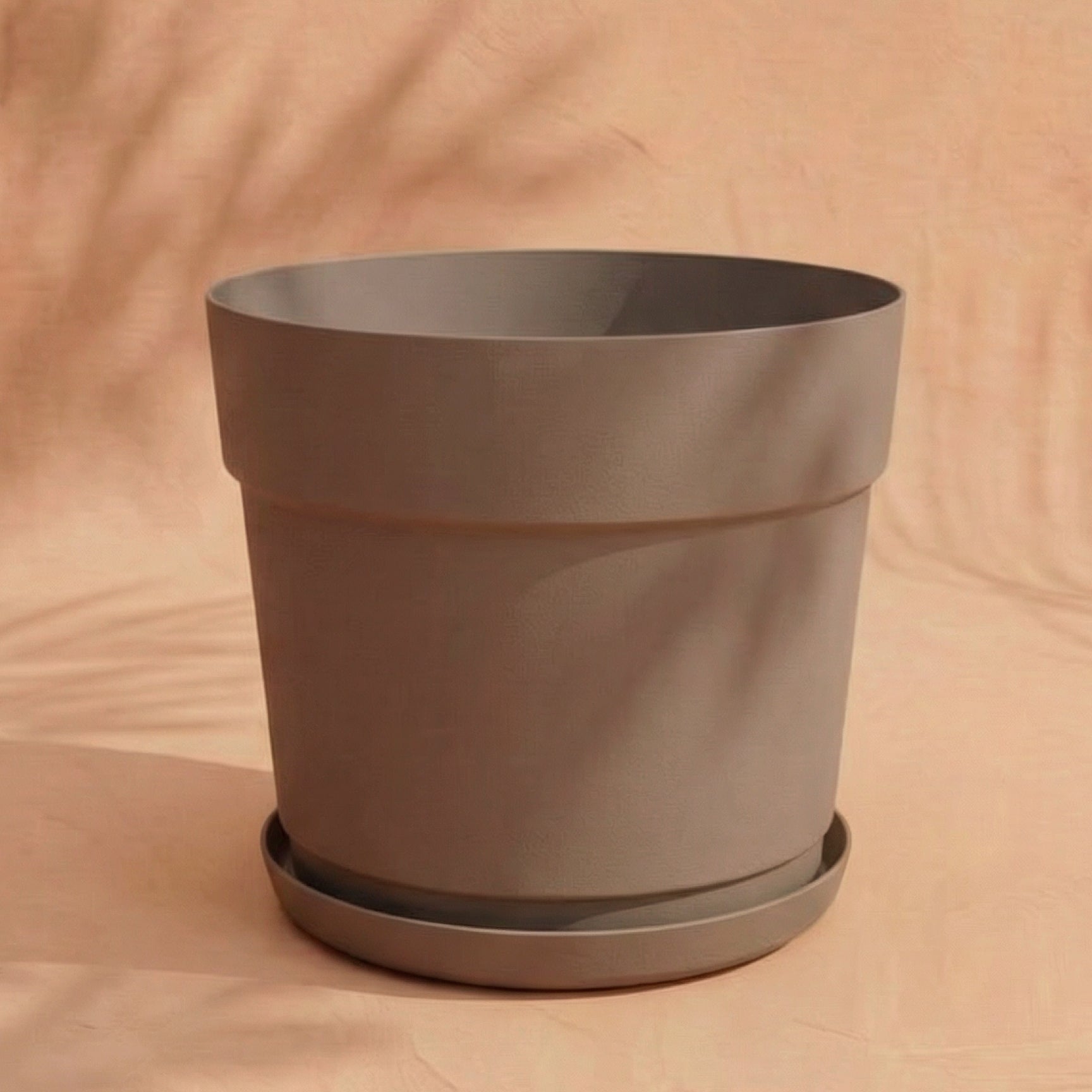 Ein neutraler Kunststoff-Pflanzkübel (Bloom | 29 cm Ø) aus recyceltem Kunststoff mit Untertasse ruht auf einer hellbraunen Oberfläche, die vor einem sanften beigen Hintergrund steht - UV-beständig und einfach elegant für jeden Raum.