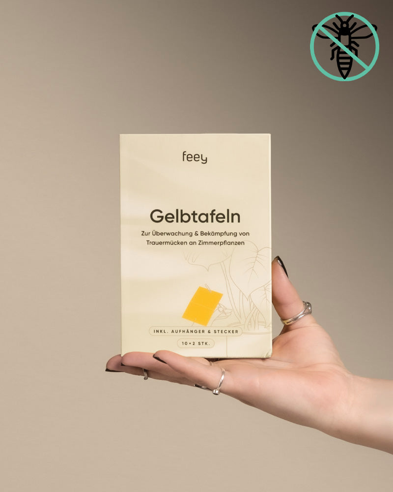 Eine Hand mit Ringen hält vor beigem Hintergrund eine Schachtel mit der Aufschrift „Gelbtafeln“. Die Schachtel dient der Überwachung und Bekämpfung von Schädlingen wie Trauermücken an Zimmerpflanzen und trägt in der oberen rechten Ecke ein durchgestrichenes Mückensymbol.