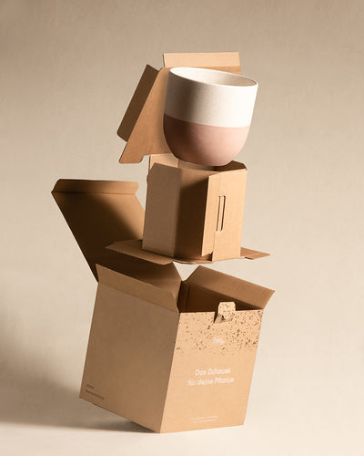 Der Keramiktopf „Variado“ aus der Keramik Topfset-Kollektion im zweifarbigen Design balanciert auf geöffneten Kartons mit der Aufschrift „Das Zuhause für deine Pflanze“ vor einem neutralen beigen Hintergrund.