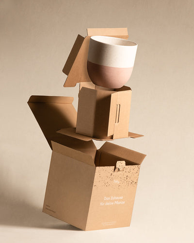 Ein schöner Keramik-Topf (Variado | 22 cm ⌀) aus der Topfkollektion Variado mit gesprenkeltem weißen Deckel und rosa-beigem Boden wird ausgepackt. Der wasserfeste Topf wird in ineinander geschachtelten Kartons vor einem hellbeigen Hintergrund präsentiert. Auf einem der Kartons steht der deutsche Text „Das Zuhause für deine Pflanze“.