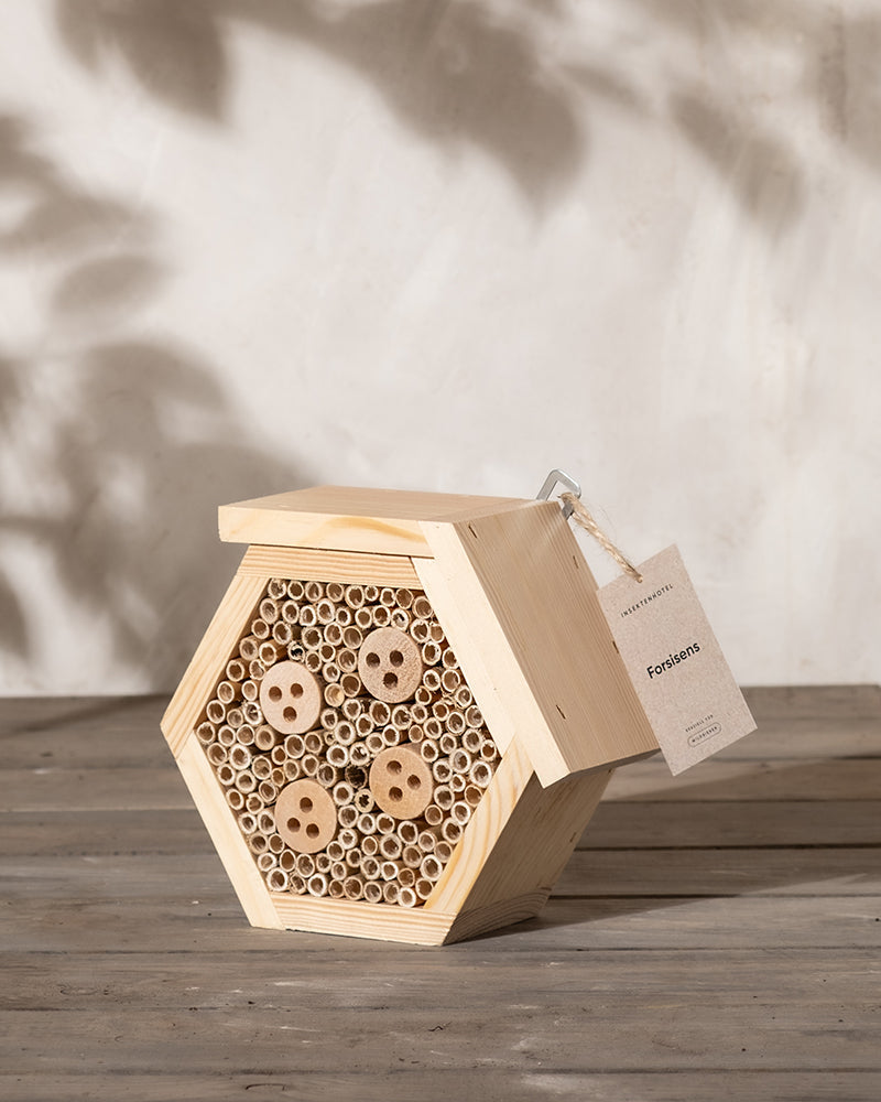 Ein kleines sechseckiges Bienenhaus aus Holz, oder Insektenhotel, mit mehreren zylindrischen Röhren und runden Löchern steht auf einer Holzoberfläche. Mit der Aufschrift „Forsisens“ ist es perfekt für Wildbienen in Ihrem Garten. Schatten von Blättern tanzen auf der beigefarbenen Wand dahinter und verleihen einen Hauch von Naturcharme.