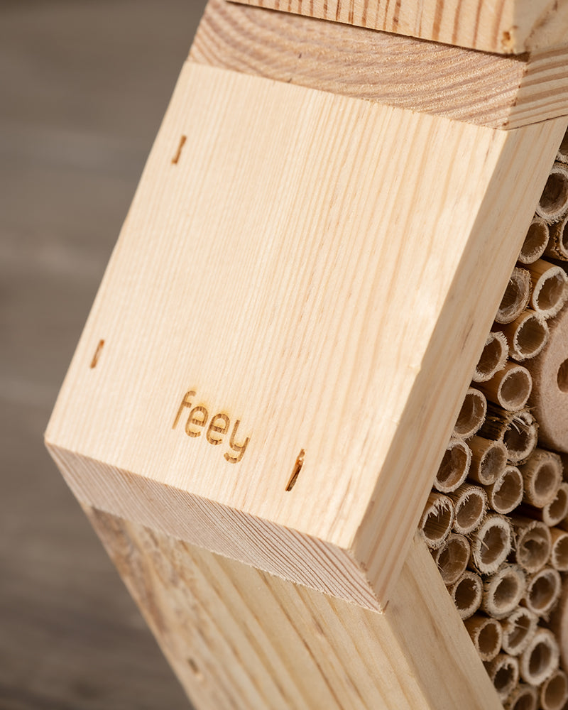 Nahaufnahme eines Insektenhotels aus hellem Holz 'Forsisens' mit kleinen zylindrischen Löchern und dem eingravierten Text "feey". Der Hintergrund ist verschwommen, was die Aufmerksamkeit auf die detaillierte Textur des Holzes und den eingravierten Text lenkt. Perfekt für jeden Garten, bietet es ein gemütliches Zuhause für Wildbienen.