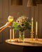Eine Hand hält ein großes Glas mit einem gelb-orangenen Getränk neben einem runden Tisch mit dem Strauß "So kunterbunt wie Du" in einer Vase, hohen Kerzenhaltern und dekorativen Kerzen, die alle warm beleuchtet sind.