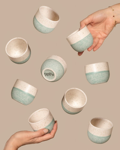 Mehrere Keramikbecher, die dem Keramik-Baby-Topfset „Variado“ (9 Stk.) mit gesprenkeltem Design in Cremeweiß und Grün ähneln, stehen auf beigem Hintergrund. Hände halten einige Becher, auf deren Unterseite „Feey“ zu sehen ist.