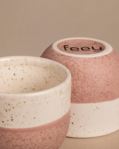 Zwei gesprenkelte Keramiktassen in Rosa-Weiß erinnern an bezaubernde Keramiktöpfe. Eine umgedrehte Tasse zeigt auf ihrem Boden „feey“ vor neutralem Hintergrund. Aus dem Keramik Baby-Topfset „Variado“ (9 Stk.).