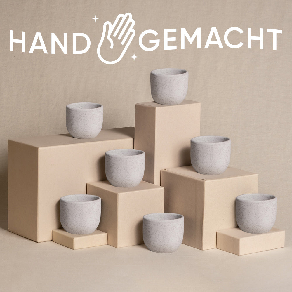 Ein Arrangement aus sieben minimalistischen Steinbechern, die auf beigefarbenen Kisten unterschiedlicher Höhe platziert sind und jeweils einen natürlichen Touch bieten. Über der Szene sind das Wort „HANDGEMACHT“ und ein Handsymbol deutlich zu sehen, was auf handgefertigte Handwerkskunst hinweist. Der Hintergrund ist ein neutraler, strukturierter Stoff.

Möchten Sie, dass ich Ihnen bei etwas anderem helfe?