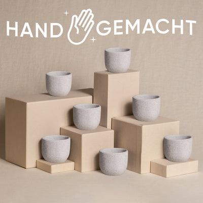 Ein Arrangement aus sieben minimalistischen Steinbechern, die auf beigefarbenen Kisten unterschiedlicher Höhe platziert sind und jeweils einen natürlichen Touch bieten. Über der Szene sind das Wort „HANDGEMACHT“ und ein Handsymbol deutlich zu sehen, was auf handgefertigte Handwerkskunst hinweist. Der Hintergrund ist ein neutraler, strukturierter Stoff.

Möchten Sie, dass ich Ihnen bei etwas anderem helfe?