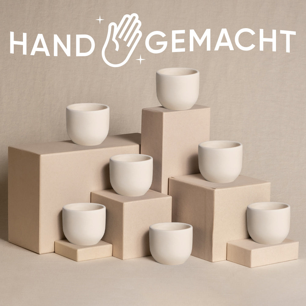 Ein Display aus sieben weißen, minimalistischen Babypflanzen-7er-Topfsets Keramik ist auf verschiedenen rechteckigen Sockeln in Beige vor einem ähnlich farbigen Hintergrund angeordnet. Das Wort „HANDGEMACHT“ mit einem stilisierten Handsymbol ist prominent über den Tassen angebracht und verleiht der Szene eine natürliche Note.
