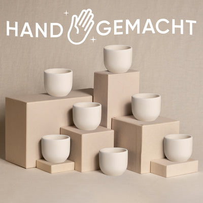 Ein Display aus sieben weißen, minimalistischen Babypflanzen-7er-Topfsets Keramik ist auf verschiedenen rechteckigen Sockeln in Beige vor einem ähnlich farbigen Hintergrund angeordnet. Das Wort „HANDGEMACHT“ mit einem stilisierten Handsymbol ist prominent über den Tassen angebracht und verleiht der Szene eine natürliche Note.