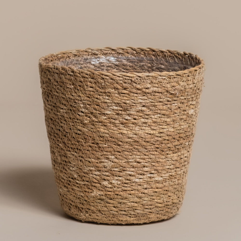 Ein runder, handgeflochtener Pflanzkorb aus Seegras (Kesat | 16 cm ⌀) in Naturbraun mit leicht konischer Form und strukturierter Oberfläche, präsentiert auf einem schlichten beigen Hintergrund.