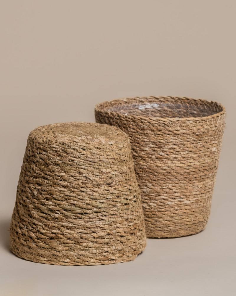 Das Pflanzkorb-Set aus Seegras (Kesat | 2× 16) besteht aus zwei strukturierten, rustikalen Körben - einem aufrechten und einem umgedrehten -, die vor einem neutralen Hintergrund stehen und jedem Raum einen Hauch von Ferienfeeling verleihen.