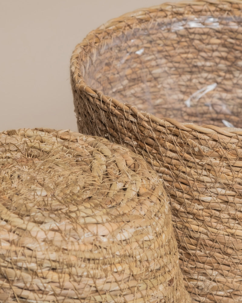 Nahaufnahme des Kesat Pflanzkorb-Sets aus Seegras (2× 16): einer stehend mit sichtbarem Kunststoffeinsatz, einer liegend, beide vor einem neutralen beigen Hintergrund für einen Hauch von Ferienfeeling.