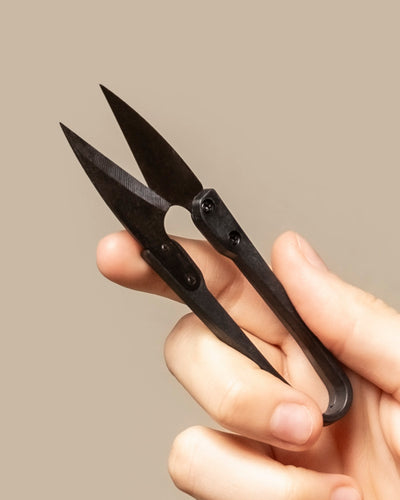 Eine Hand hält vor einem schlichten beigen Hintergrund eine kleine Pflanzenschere mit scharfen, spitzen Klingen, ideal zum Nähen, Basteln oder Schneiden von Stecklingen.