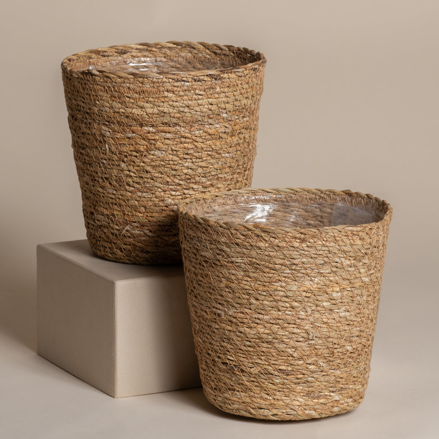 Zwei Pflanzkorb-Set aus Seegras (Kesat | 2×16) mit Klarsichtfolie - einer auf einem beigen Block, einer auf dem Boden. Das natürliche Design bringt Ferienfeeling in jeden Raum, hier vor einem schlichten beigen Hintergrund.