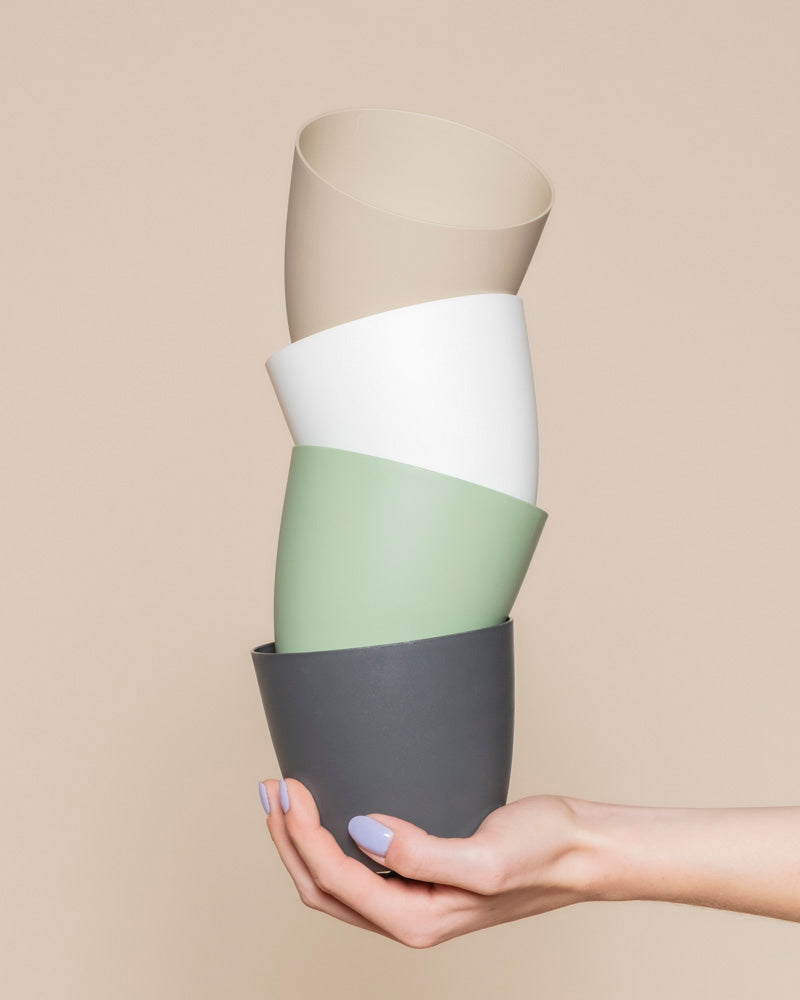 Eine Hand mit lavendelfarbenem Nagellack hält vier gestapelte Kunststoff-Töpfe (Kiki | 11 cm Ø) in den Farben grau, grün, weiß und beige-matt, die sich perfekt für Ihre Pflänzchen und Ihr Zuhause eignen, vor einem beigen Hintergrund.