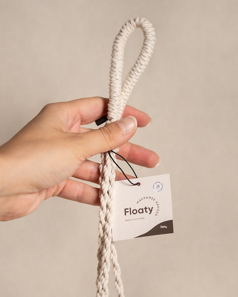 Eine Hand hält einen cremefarbenen Seilanhänger mit Schlaufe, der mit dem Etikett „Makramee-Hänger Floaty (3er)“ beschriftet ist und die Form elegant widerspiegelt. Das Etikett zeigt die Produktdetails auf neutralem Hintergrund und unterstreicht so die Eleganz des Anhängers.