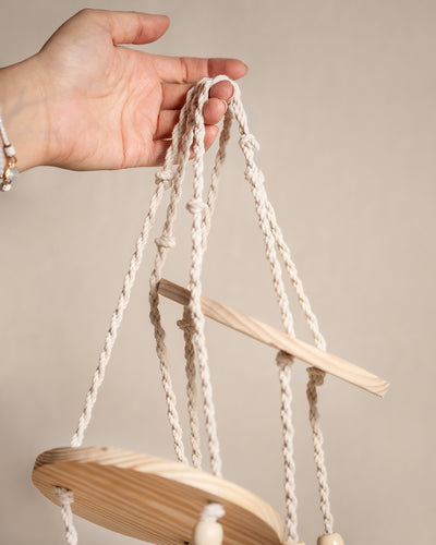 Eine Hand hält ein geknotetes weißes Seil, das mit runden Holzplattformen verbunden ist, den Elementen des Makramee-Hängers Floaty (3er). Der neutrale Hintergrund unterstreicht die natürliche Ästhetik dieser Hängedekoration für Jungpflanzen.