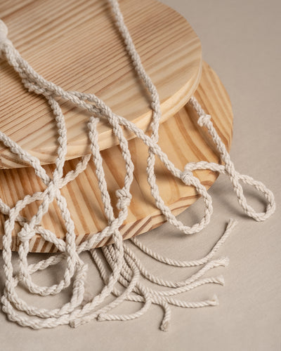 Nahaufnahme des Makramee-Hängers „Floaty“ (2er), bestehend aus geflochtenen weißen Seilen, die auf runden, unbehandelten Holzbrettern mit natürlicher Maserung verflochten sind. Die Seile kreuzen sich auf einer beigen Stoffoberfläche, ideal, um kleine Räume mit Pflanzendekor aufzuwerten.