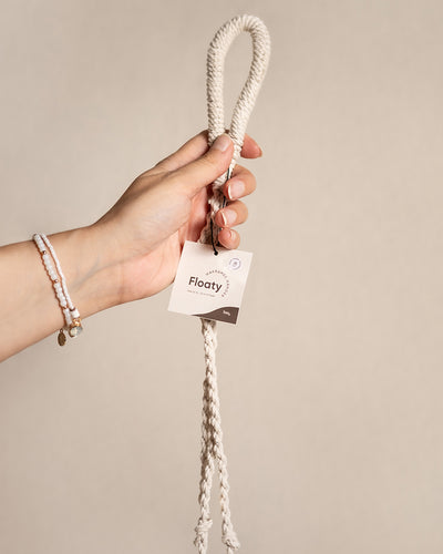 Eine Hand hält ein cremefarbenes geflochtenes Seil mit einer Schlaufe, beschriftet mit „Makramee-Hänger 'Floaty' (2er)“, perfekt zum Dekorieren kleiner Räume. Vor einem neutralen Hintergrund verleiht es jedem Makramee-Regal Charme.