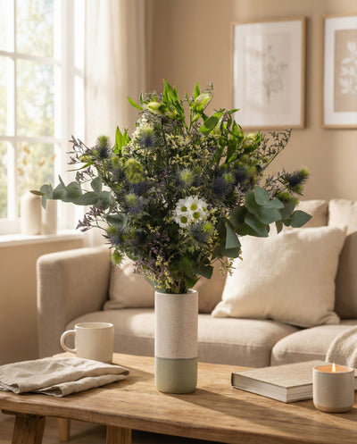 Der Blumenstrauß "Nebelmeer" aus zarten Blüten schmückt den Couchtisch im gemütlichen Wohnzimmer. Neben ihm stehen eine Tasse, ein Buch, ein gefaltetes Tuch und eine brennende Kerze. Sanfte Morgensonne fällt durchs Fenster.