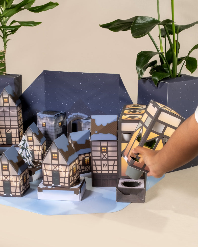Eine Hand öffnet einen Pflanzen-Adventskalender aus Papier in Form eines Winterdorfs, wobei jedes Gebäude in festlichen Farben geschmückt und fortlaufend nummeriert ist. Im Hintergrund sind echte Pflanzen kunstvoll arrangiert, um die Szene auf einer flachen Oberfläche zu verschönern.