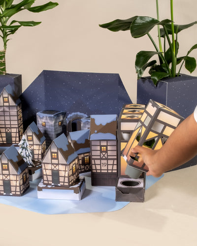 Eine Hand öffnet einen Pflanzen-Adventskalender aus Papier in Form eines Winterdorfs, wobei jedes Gebäude in festlichen Farben geschmückt und fortlaufend nummeriert ist. Im Hintergrund sind echte Pflanzen kunstvoll arrangiert, um die Szene auf einer flachen Oberfläche zu verschönern.