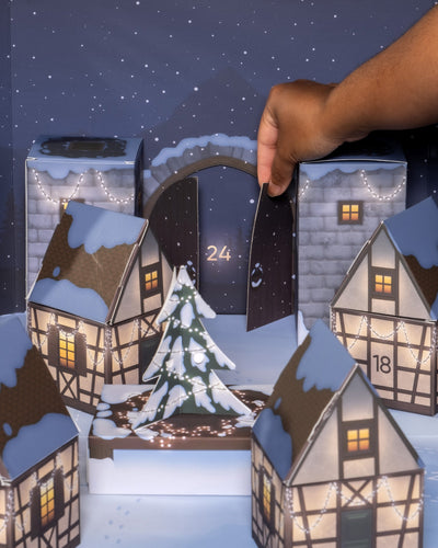 Eine Hand öffnet Türchen 24 des Pflanzen-Adventskalenders. Darin ist eine Schneelandschaft mit kleinen, schneebedeckten Häusern und Bäumen vor der Kulisse einer sternenklaren Nacht zu sehen, die von festlichen Lichterketten wunderschön beleuchtet wird.