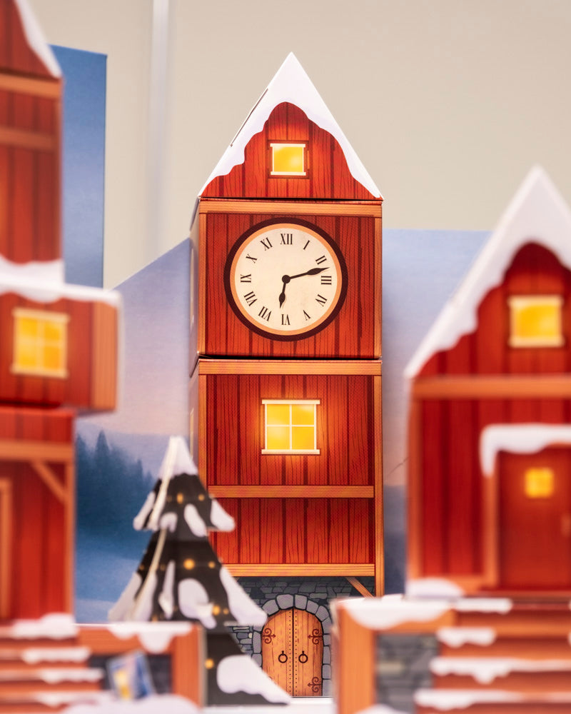 Ein Modell des Pflanzen-Adventskalenders (Dschungel-Version) mit einem roten hölzernen Uhrenturm mit verschneitem Dach, leuchtenden Fenstern und einem runden Ziffernblatt um 10:10 Uhr. Schnee bedeckt die umliegenden Gebäude, Pflanzgefäße und einen Baum und sorgt für ein gemütliches Winterdorfgefühl.