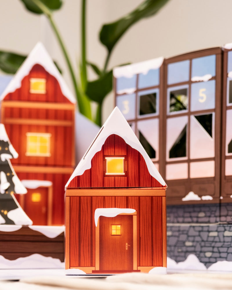 Eine Nahaufnahme von dekorativen Papierhäusern mit schneebedeckten Dächern und leuchtenden Fenstern, vor einem Pflanzen-Adventskalender (Dschungel-Version) und üppig grünen Zimmerpflanzen im Hintergrund.