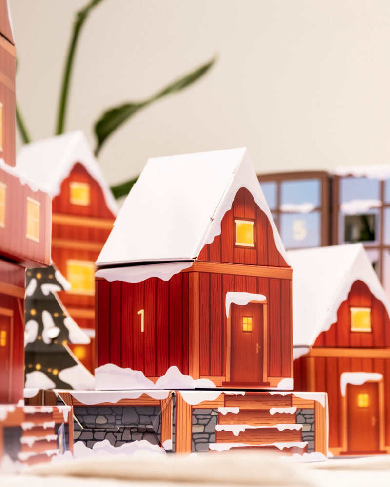 Eine Nahaufnahme des Pflanzen-Adventskalenders (Dschungel-Version), ein festliches Holzhäuschen mit der Nummer 1, einem verschneiten Dach und leuchtenden Fenstern, umgeben von zauberhaften Schachteln, in denen sich kleine Zimmerpflanzen oder Blumentöpfe verstecken lassen.