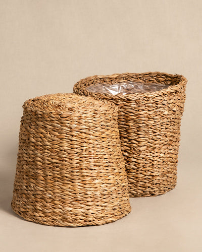 Vor einem neutralen Hintergrund sind zwei geflochtene Weidenkörbe zu sehen. Der Korb links liegt auf der Seite und gibt den Blick auf sein Inneres frei. Beim aufrecht stehenden Pflanzenkorb aus Seegras (Kesat | 22 cm ⌀) rechts ist eine transparente Kunststoffverkleidung im Inneren sichtbar. Beide haben eine strukturierte, natürliche Optik, die an eine sommerliche Oase erinnert.