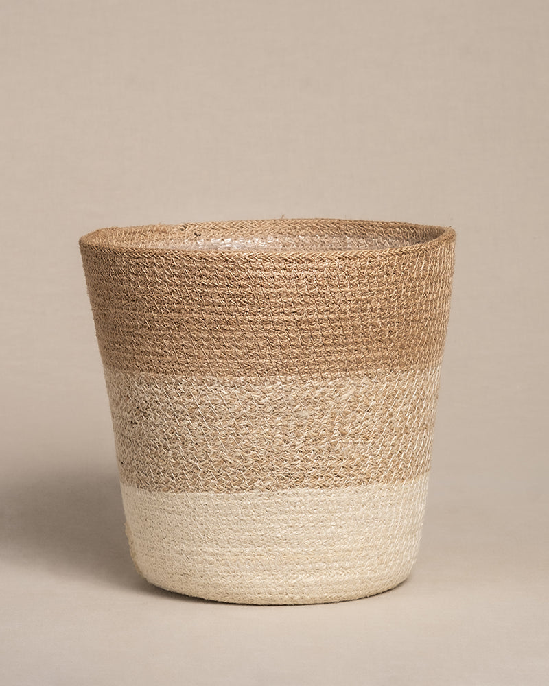 Der Pflanzenkorb aus Jute (Lapis | 22 cm Ø) hat ein zweifarbiges Design mit breiter Oberseite und schmaler Unterseite. Der obere Teil ist naturbraun, während der untere Teil heller beige ist. Dieser handgefertigte Korb verleiht jedem Raum Charme und kommt vor neutralen Hintergründen schön zur Geltung.