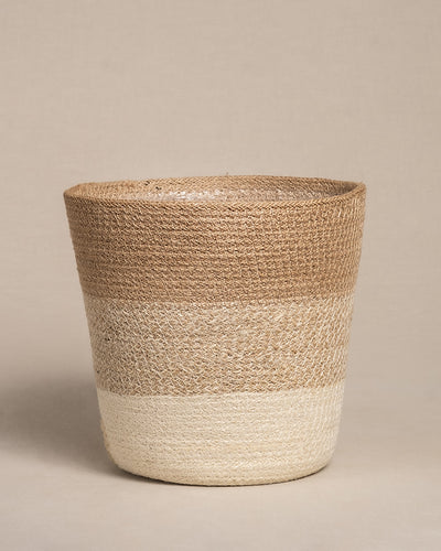Der Pflanzenkorb aus Jute (Lapis | 22 cm Ø) hat ein zweifarbiges Design mit breiter Oberseite und schmaler Unterseite. Der obere Teil ist naturbraun, während der untere Teil heller beige ist. Dieser handgefertigte Korb verleiht jedem Raum Charme und kommt vor neutralen Hintergründen schön zur Geltung.